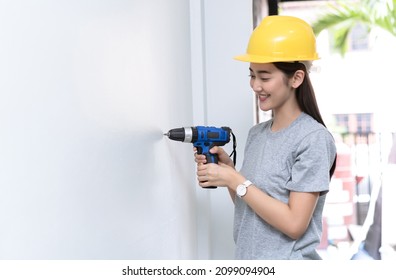 Happy Young Asian Woman Builder Drilling A Hole In A New White Wall In New House With A Battery Operated Hand Drill. Adult Making DIY Workshop. Young Female Repairing Home. Moving In New House Concept