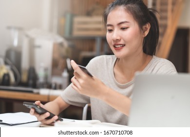 Happy young asian woman buying stuff from online retail and paying bills via banking application. Girl looking at credit card in her hand and fill data to mobile phone to complete purchese order..