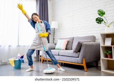 Happy young Asian woman cleaning her home, singing at mop like at microphone and having fun, free space. 