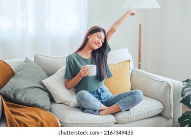 Happy young asian woman drinking coffee relaxing on sofa at home. Smiling female enjoying resting sitting on couch in modern living room.