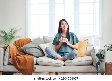 Happy young asian woman drinking coffee relaxing on sofa at home. Smiling female enjoying resting sitting on couch in modern living room. - Powered by Shutterstock