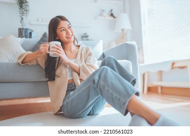 Happy young asian woman drinking coffee relaxing on the rug beside to the sofa at home. Smiling female enjoying resting sitting on couch in modern living room. - Powered by Shutterstock