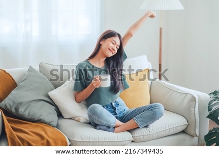 Happy young asian woman drinking coffee relaxing on sofa at home. Smiling female enjoying resting sitting on couch in modern living room.