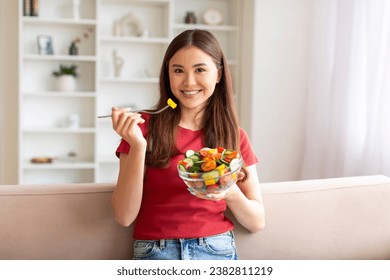 Happy young asian woman eating vegetable salad while sitting on couch in living room, smiling beautiful lady having healthy lunch, enjoying fresh veggies, relaxing in home interior, copy space - Powered by Shutterstock