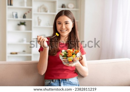Happy young asian woman eating vegetable salad while sitting on couch in living room, smiling beautiful lady having healthy lunch, enjoying fresh veggies, relaxing in home interior, copy space