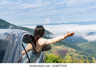 Happy young Asian woman enjoying sitting in a car watching a beautiful mountain view, hand greeting. Driving road trip on vacation and adventure concept