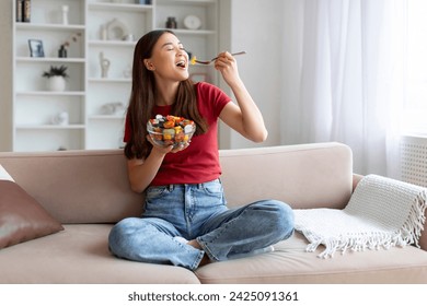 Happy young asian woman enjoying fresh salad bowl at home, beautiful korean lady laughing while eating healthy food, sitting on comfortable sofa in well-lit, tastefully decorated living room - Powered by Shutterstock