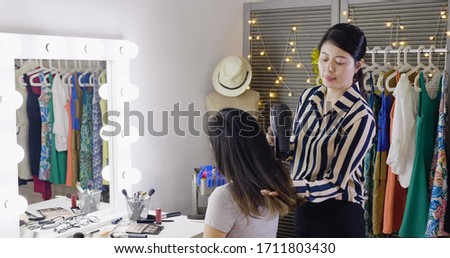 Happy young asian woman entertainer and hairdresser with fan making hot styling at dressing place. girl celebrity sitting at vanity table in makeup room while stylist using hair drying blowing.