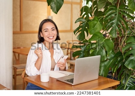 Happy young asian woman feel success, winning and celebrating, sitting with laptop and smartphone in cafe and triumphing, shouting yes.