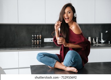 Happy Young Asian Woman In Headphones Eating Dessert With A Spoon From A Jar In A Kitchen