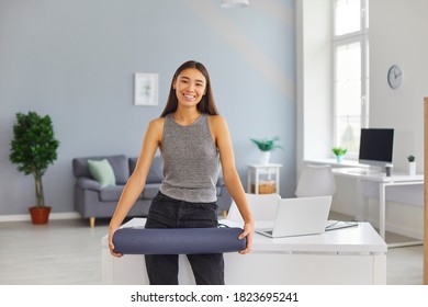 Happy Young Asian Woman Holding Yoga Mat Standing Near Office Desk. Smiling Employee Taking Break From Work In Cozy Modern Workplace. Concept Of Workplace Health And Corporate Wellness Programs