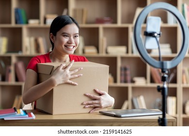 Happy Young Asian Woman Hugging Cardboard Box And Look At Smartphone Webcam In Living Room Interior. Unboxing At Home, Online Shopping, Blog, Human Emotions And New Normal, Social Distance And Tech