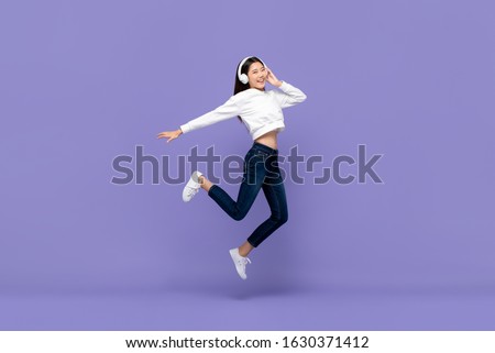 Happy young Asian woman jumping and listening to music on headphones isolated on purple background