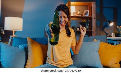 Happy young asian woman looking at camera enjoy night party event online with friends toast drink beer via video call online in living room at home, Stay at home quarantine, Social distancing concept.