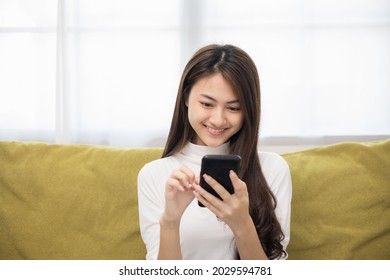 Happy young asian woman looking at screen of smartphone sitting on sofa in living room she shopping online. She very happy. Teenage girl reading news from mobile phone feeling excited.