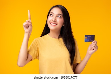 Happy young Asian woman pointing up to copy space and looking at copy space with smile face holding credit card Pretty girl act like a satisfied product use for advertisement with yellow background