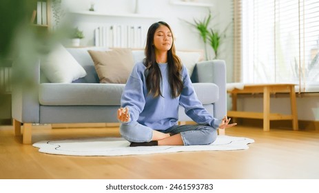 Happy young Asian woman practicing yoga and meditation at home sitting on floor in living room in lotus position and relaxing with closed eyes. Mindful meditation and wellbeing concept - Powered by Shutterstock