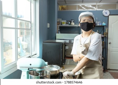 Happy Young Asian Woman With Protective Face Mask Is Smiling, Holding Kitchen Tools While Cooking, Baking Homemade Bakery Cake, Food In Home Kitchen When Home Quarantine In Coronavirus Disease Spread.