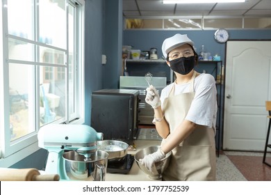 Happy Young Asian Woman With Protective Face Mask Is Smiling, Holding Kitchen Tools While Cooking, Baking Homemade Bakery Cake, Food In Home Kitchen When Home Quarantine In COVID-19 Disease Spread.