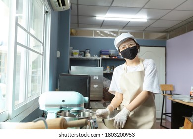 Happy Young Asian Woman With Protective Face Mask Is Smiling, Holding Kitchen Tools While Cooking, Baking Homemade Bakery Cake, Food In Home Kitchen When Home Quarantine In Coronavirus Disease Spread.