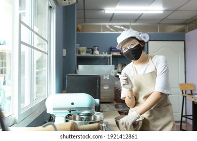 Happy Young Asian Woman With Protective Face Mask Is Smiling, Holding Kitchen Tools While Cooking, Baking Homemade Bakery Cake, Food In Home Kitchen When Home Quarantine In COVID-19 Or Coronavirus.