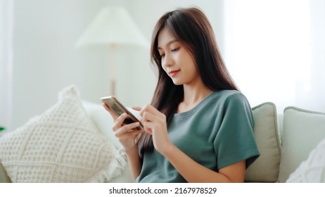 Happy young asian woman relax on comfortable couch at home texting messaging on smartphone, smiling girl use cellphone chatting, browse wireless internet on gadget, shopping online from home - Powered by Shutterstock