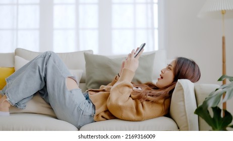 Happy Young Asian Woman Relax On Comfortable Couch At Home Texting Messaging On Smartphone, Smiling Girl Use Cellphone, Browse Wireless Internet On Gadget, Shopping Online From Home