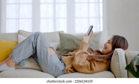 Happy Young Asian Woman Relax On Comfortable Couch At Home Texting Messaging On Smartphone, Smiling Girl Use Cellphone, Browse Wireless Internet On Gadget, Shopping Online From Home