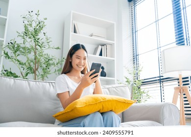 Happy young Asian woman relax sitting on couch using cell phone, smiling lady laughing holding smartphone, looking at cellphone enjoying doing online ecommerce shopping in mobile apps - Powered by Shutterstock