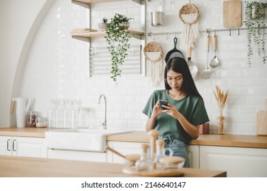 Happy young asian woman relaxing at home she is standing at counter kitchen and using mobile smartphone
