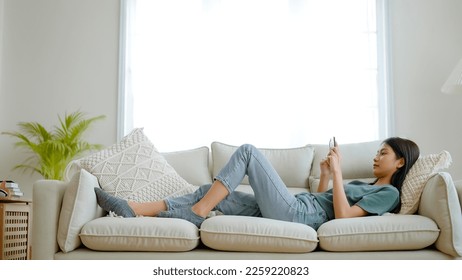 Happy young asian woman relaxing at home. Female is lying down on sofa and using mobile smartphone