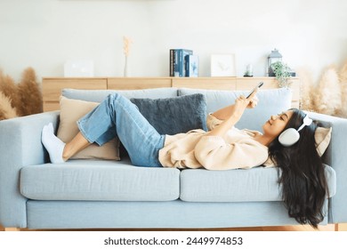 Happy young asian woman relaxing at home. Female smile sitting on sofa and holding mobile smartphone. Girl using video call to friend. Listening to music - Powered by Shutterstock