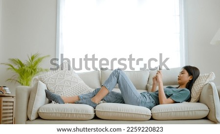 Happy young asian woman relaxing at home. Female is lying down on sofa and using mobile smartphone