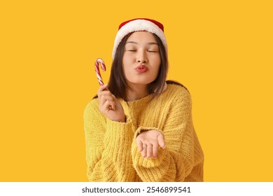 Happy young Asian woman in Santa hat with candy cane blowing kiss on yellow background - Powered by Shutterstock