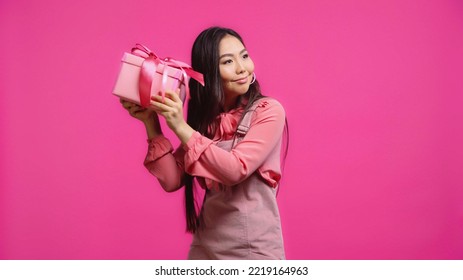 Happy Young Asian Woman Shaking Wrapped Present Isolated On Pink 