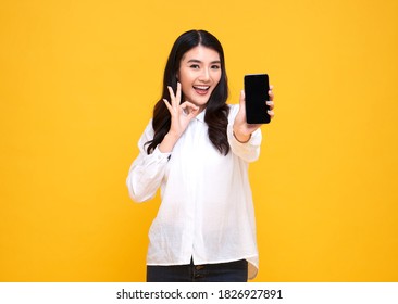 Happy Young Asian Woman Showing At Blank Screen Mobile Phone And Hand Gesture Ok Isolated Over Yellow Background.