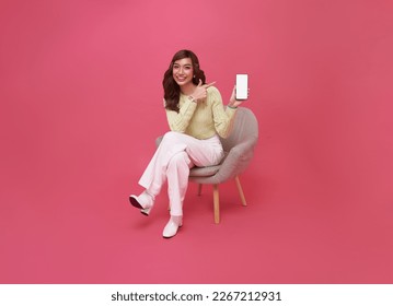 Happy young Asian woman showing empty screen mobile phone. While her sitting on chair isolated on pink studio background.