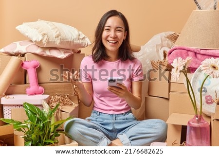 Happy young Asian woman sits crossed legs uses smartphone packs up for relocation in new apartment buys furniture online for her house surrounded by cardboard boxes full of stuff. Moving day