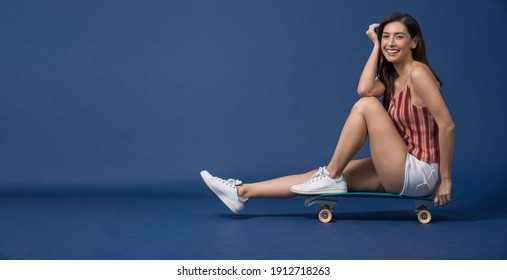 Happy Young Asian Woman Sitting And Holding Surfskate Or Skateboard On Blue Color Background, Exercise And Sport, Trendy And Extream Sports Concept