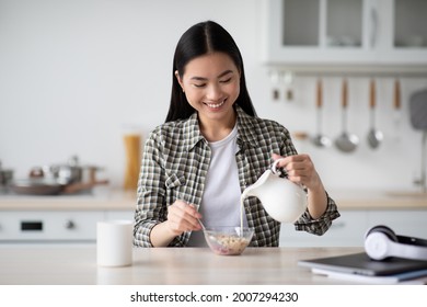 Happy young asian woman sitting at white cozy kitchen eating healthy oatmeal, pouring milk at glass bowl with granola, enjoying her breakfast, copy space. Healthy lifestyle, diet, nutrition concept - Powered by Shutterstock