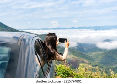 Happy young Asian woman sitting in a car while take a photo and watching a beautiful mountain view, hand greeting. Driving road trip on vacation and adventure concept. Copy space - Powered by Shutterstock