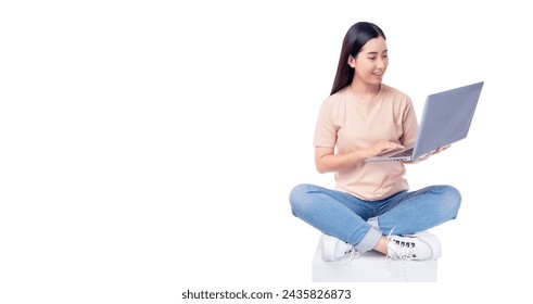 Happy young asian woman sitting on white chair with legs crossed and using laptop computer on light pink background copy space Full body young smiling happy female wearing t shirt and jeans lifestyle