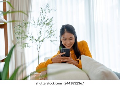 Happy young Asian woman sitting on sofa holding mobile phone using cellphone technology doing ecommerce shopping, buying online, texting messages relaxing on couch in cozy living room at home - Powered by Shutterstock