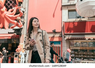 Happy Young Asian Woman With Smartphone Standing In Street In Dotonbori Area. Office Lady Holding Mobile Phone And Open Map App Looking Aside Searching Way In Osaka Japan. Huge Octopus Sign In Back.