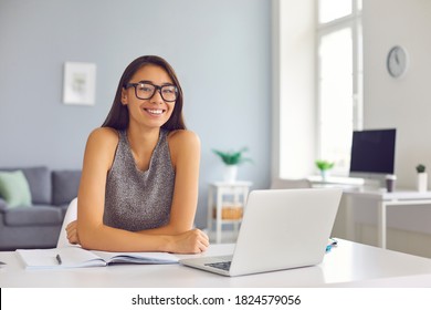 Happy Young Asian Woman In Stylish Glasses Sitting At Desk With Open Laptop And Notebook Looking At Camera. Successful Smiling Student Girl Staying At Home Using Online Learning Tools And Platforms