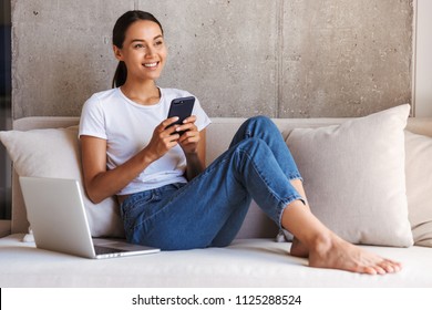 Happy Young Asian Woman Using Mobile Phone While Sitting On A Couch At Home With Laptop Computer