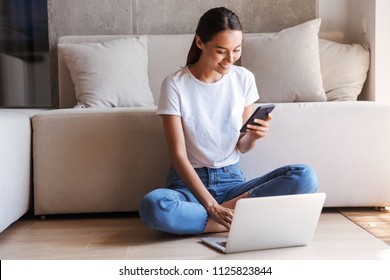 Happy Young Asian Woman Using Mobile Phone While Sitting At A Couch At Home With Laptop Computer