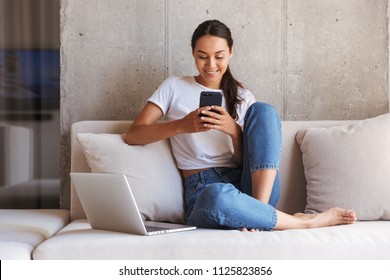 Happy young asian woman using mobile phone while sitting a couch at home with laptop computer - Shutterstock ID 1125823856