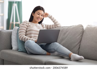 Happy Young Asian Woman Watching Movies Online On Laptop At Home, Cheerful Korean Female Relaxing On Comfortable Couch With Computer, Using Modern Technologies For Leisure, Enjoying Domestic Pastime