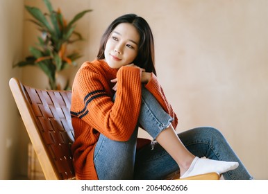 Happy young Asian woman wearing a sweater sitting on wooden chair and looking outside in living room at home on a bright winter morning. Concept woman lifestyle and winter. Autumn, winter season.